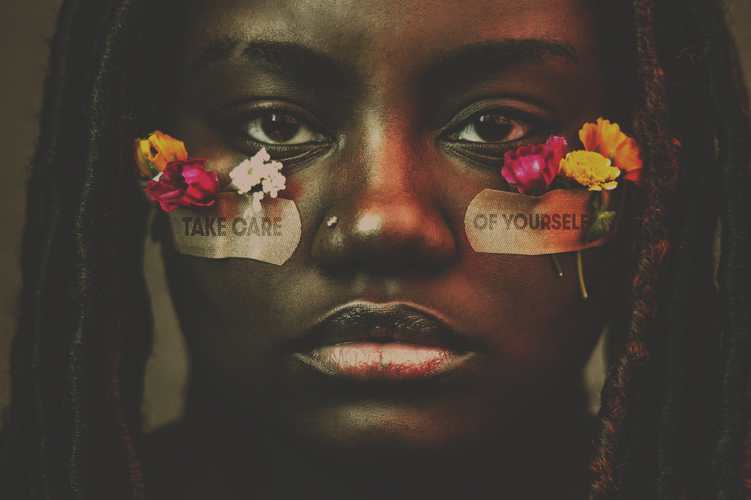 Young Black woman facing camera with small flowers fixed to her face by bandaids that have 'Take Care of Yourself' written on them